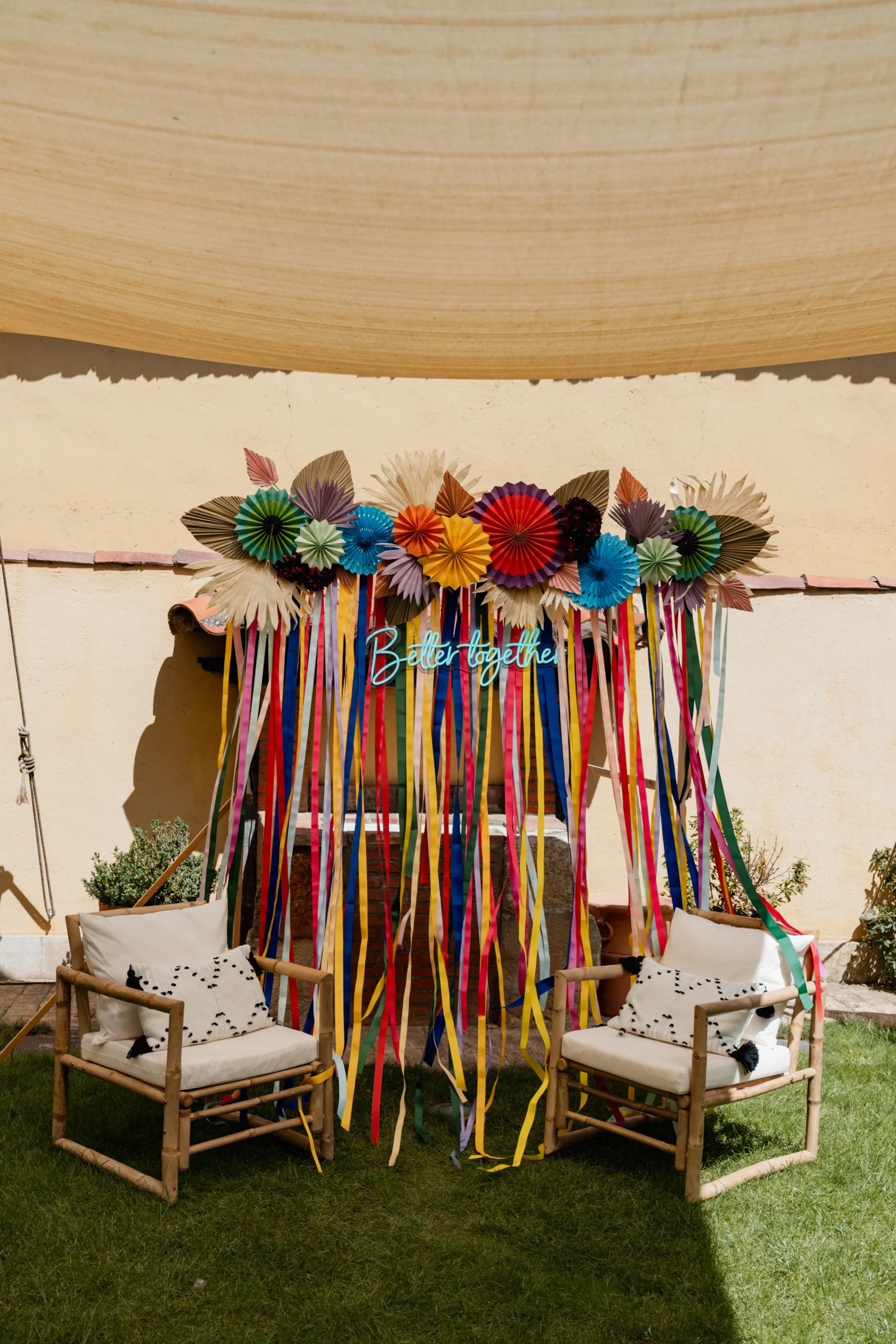 Photocall alegre y colorido con abanicos de papel y cintas de colores, acompañado de dos sillones de bambú con cojines sobre césped, ideal para fotos originales en celebraciones y bodas.