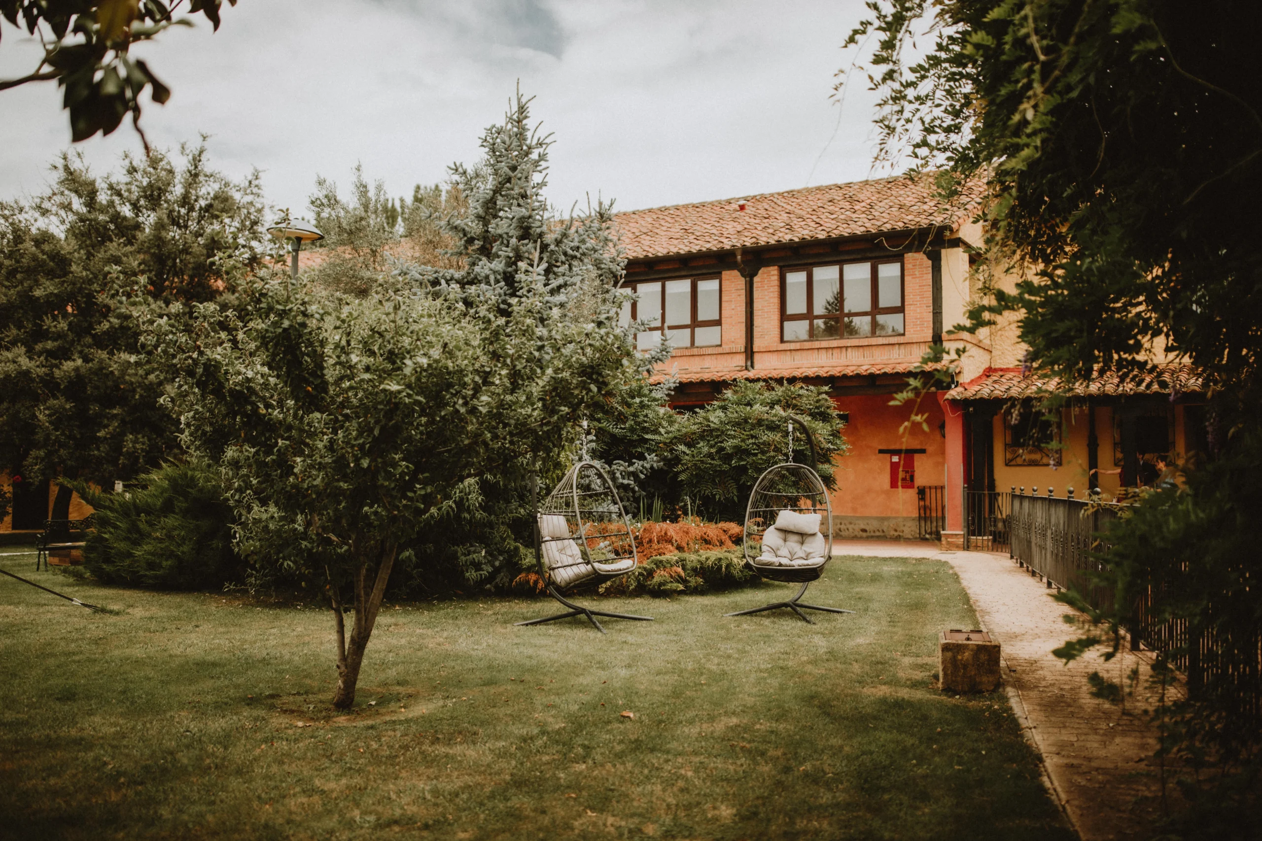 Jardín amplio y cuidado con columpios tipo huevo y árboles floridos, junto a una casa rural, ideal para momentos de relax o fotografías románticas durante celebraciones y bodas.
